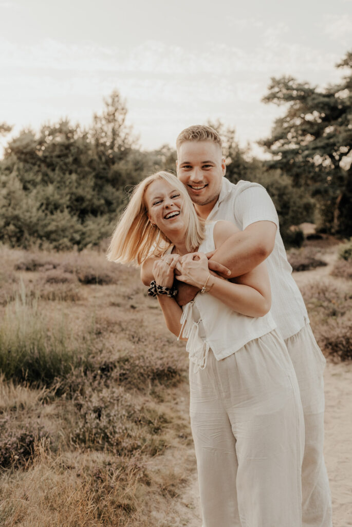 Paar beim Fotoshooting in der Heide, beide tragen helle Klamotten und lachen in die Kamera