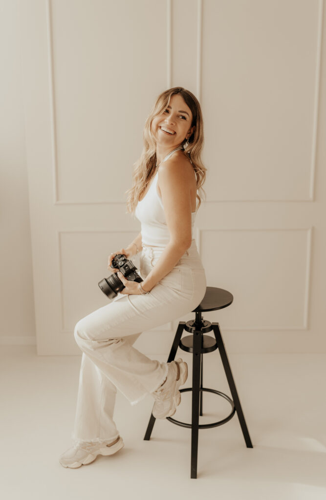 Fotografin Vicky mit Kamera in der Hand, sitzt auf Hocker und lacht in die Kamera
